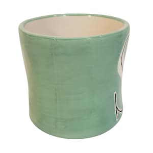 May include: A light green ceramic mug with a white interior. The mug has a curved shape and a design on the side. The mug is suitable for holding hot or cold beverages.