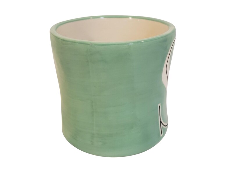 May include: A light green ceramic mug with a white interior. The mug has a curved shape and a design on the side. The mug is suitable for holding hot or cold beverages.