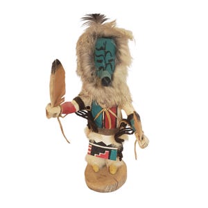 May include: A handcrafted kachina doll featuring a turquoise face with black markings, surrounded by light brown fur. The figure wears a painted red, white, and black garment, holds a brown feather in one hand, and stands on a circular wooden base.