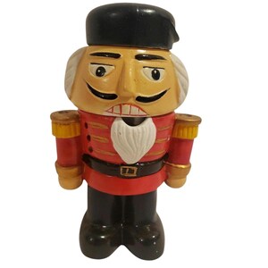 Puede incluir: Figura decorativa de cascanueces con forma de soldado. La figura tiene un sombrero negro, una cara color canela con barba blanca y un uniforme rojo con detalles dorados. El cascanueces está de pie.