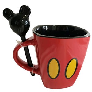 May include: A red ceramic mug with a black interior and two yellow oval shapes on the front. The mug features a handle with an attached black spoon that has a handle shaped like the silhouette of a mouse head with two round ears.