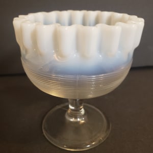 May include: A vintage, opalescent glass dessert dish. The bowl has a ruffled rim and sits atop a clear, ribbed base and stem. The glass has a milky white color with a hint of blue, and the foot is 3 inches in diameter.
