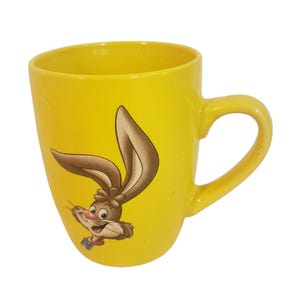 Puede incluir: Taza de cerámica amarilla con un diseño de conejo de dibujos animados. La taza tiene un asa curva y una superficie lisa. El conejo tiene orejas marrones y una cara sonriente. Una taza colorida para bebidas.