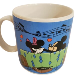 K&ouml;nnte beinhalten: Keramik-Kaffeetasse mit Mickey und Minnie Mouse in einer romantischen Szene. Die Tasse hat einen blauen Hintergrund mit Noten und Herzen. Der Griff und das Innere sind wei&szlig;. Das Design beinhaltet einen Plattenspieler.