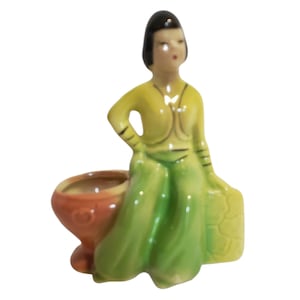 Op de afbeelding: Een keramieken beeldje van een persoon met donker haar in een geel shirt en een groene broek, zittend naast een kleine, ronde, oranje kom. De figuur heeft een hand in de zij en staat naast een gestructureerde, gele rechthoekige basis.