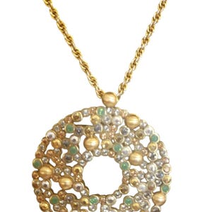May include: Gold-coloured necklace with a circular pendant decorated with gold, silver, and green beads and crystals. The pendant has a central hole and hangs from a twisted rope chain.