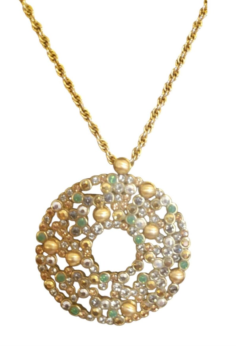 May include: Gold-coloured necklace with a circular pendant decorated with gold, silver, and green beads and crystals. The pendant has a central hole and hangs from a twisted rope chain.