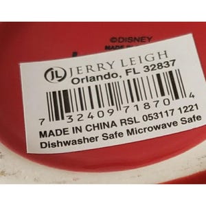 May include: A red ceramic base with a white rectangular product label featuring the text JERRY LEIGH Orlando, FL 32837, a barcode, the text MADE IN CHINA RSL 053117 1221, and the text Dishwasher Safe Microwave Safe, with &copy;DISNEY printed above.