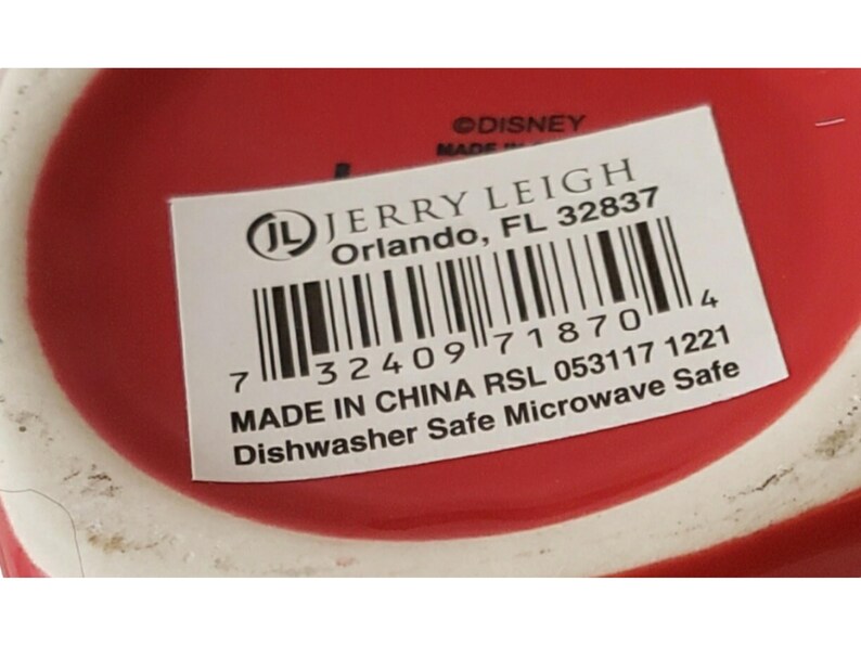 May include: A red ceramic base with a white rectangular product label featuring the text JERRY LEIGH Orlando, FL 32837, a barcode, the text MADE IN CHINA RSL 053117 1221, and the text Dishwasher Safe Microwave Safe, with &copy;DISNEY printed above.