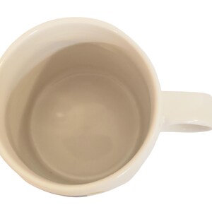 May include: Cream-colored ceramic mug with a rounded body and a rectangular handle. The interior of the mug is a light beige color. The mug is empty and viewed from above, showing the inside and handle.