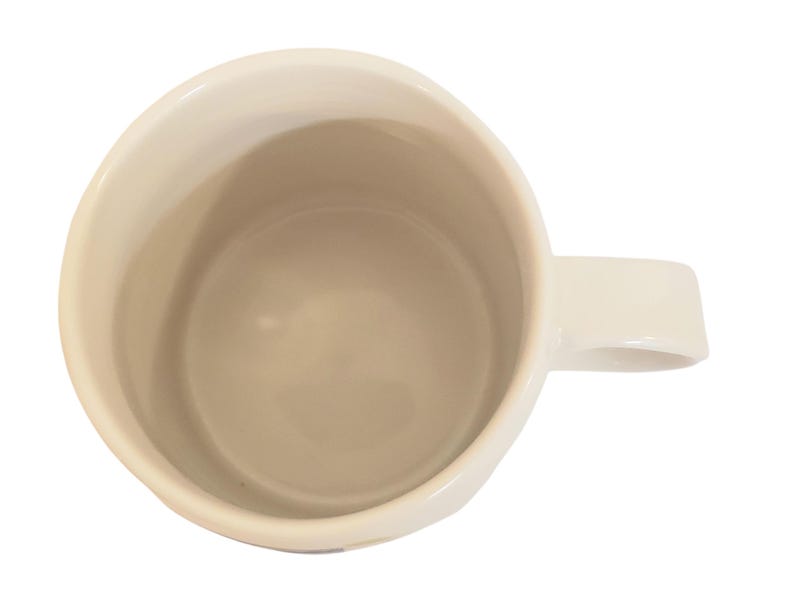 May include: Cream-colored ceramic mug with a rounded body and a rectangular handle. The interior of the mug is a light beige color. The mug is empty and viewed from above, showing the inside and handle.
