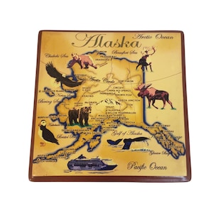 Puede incluir: Azulejo decorativo cuadrado de estilo vintage con un mapa de Alaska. El mapa incluye ilustraciones de animales salvajes como osos, alces y águilas, junto con el texto "Alaska", "Arctic Ocean" y "Pacific Ocean". El azulejo tiene un borde marrón y un fondo sepia.