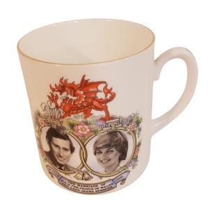 May include: A cream-colored ceramic mug featuring a commemorative design for the 1981 marriage of the Prince of Wales and Lady Diana Spencer. The mug displays a red dragon, floral motifs, and portraits of the couple with the text: THE MARRIAGE OF H.R.H THE PRINCE OF WALES AND LADY DIANA SPENCER.