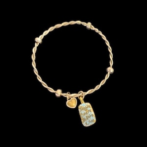 Puede incluir: Una pulsera dorada con un dise&ntilde;o trenzado. La pulsera presenta un peque&ntilde;o dije de coraz&oacute;n dorado y un dije rectangular con las palabras "Imagine, Create, Inspire, Live" en letra cursiva. El fondo es negro.