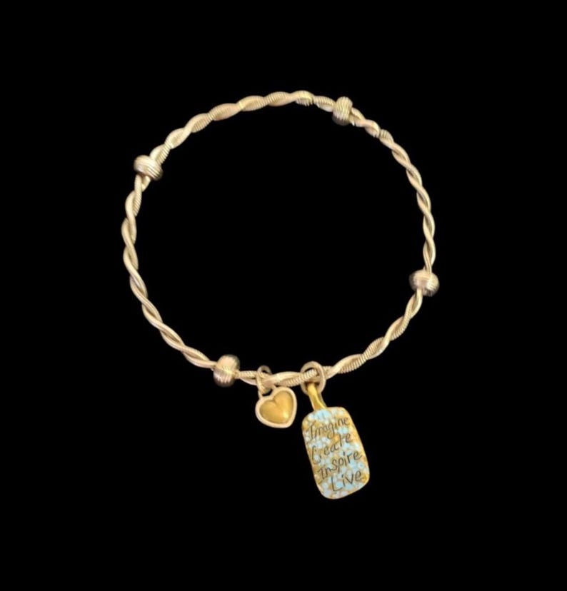 Puede incluir: Una pulsera dorada con un dise&ntilde;o trenzado. La pulsera presenta un peque&ntilde;o dije de coraz&oacute;n dorado y un dije rectangular con las palabras "Imagine, Create, Inspire, Live" en letra cursiva. El fondo es negro.