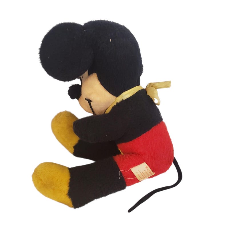 Op de afbeelding: Vintage Mickey Mouse pluche speelgoed. Het pluche heeft zwarte vacht, een gele strik en een rode broek. Het speelgoed zit in een zittende positie met gele handen en voeten. Een klein label is aan de zijkant bevestigd.