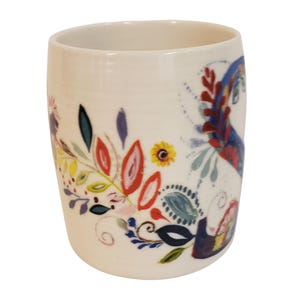 May include: A white ceramic mug with a colorful floral design. The design includes red, yellow, blue, and green leaves and flowers. The mug has a cylindrical shape and a smooth surface.