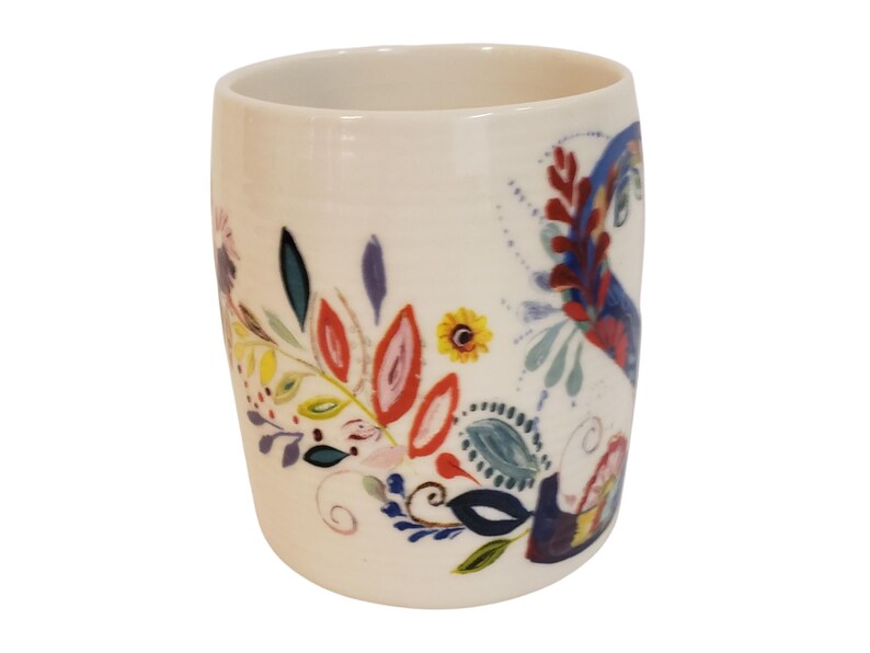 May include: A white ceramic mug with a colorful floral design. The design includes red, yellow, blue, and green leaves and flowers. The mug has a cylindrical shape and a smooth surface.