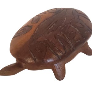Puede incluir: Una figura de tortuga de madera tallada con un acabado suave y pulido en una paleta de colores marrón bicolor. El caparazón muestra intrincados patrones geométricos empotrados grabados en la superficie, mientras que la cabeza y las patas están esculpidas.