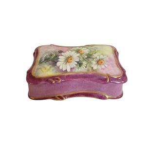 May include: A rectangular porcelain trinket box with a wavy, scalloped edge. The lid features a hand-painted floral design of white daisies with yellow centers against a soft pink and green background. The base is a deep purple with gold trim.
