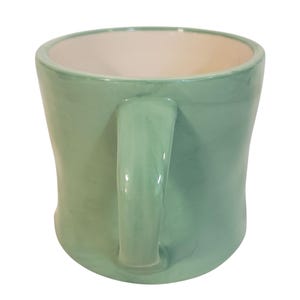 May include: A handmade ceramic mug in a soft, muted green color. The mug has a rounded shape with a sturdy handle and a contrasting off-white interior. This unique mug is perfect for enjoying your favorite hot beverages.