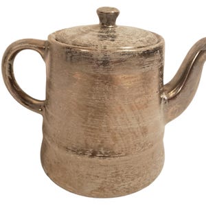 May include: A metallic teapot with a distressed, brushed bronze finish. The teapot features a cylindrical body, a curved handle on the left side, a curved spout on the right side, and a matching lid with a small knob handle on top.