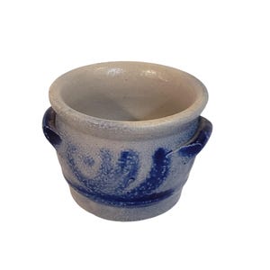 May include: A small, round, light gray ceramic crock with a speckled texture and a wide, open top. The vessel features two small, curved side handles and is decorated with a bold, hand-painted cobalt blue swirl pattern encircling the exterior.