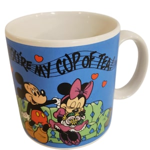 K&ouml;nnte beinhalten: Eine Keramik-Tasse mit wei&szlig;em Innenraum und Henkel, die Micky und Minnie Maus auf blauem Hintergrund zeigt. Die Tasse tr&auml;gt den Text "You're my cup of tea" mit Noten und Herz-Akzenten. Die Figuren sind in ihrem klassischen Cartoon-Stil gehalten.