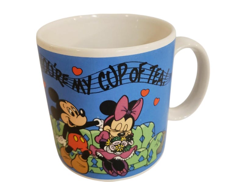 K&ouml;nnte beinhalten: Eine Keramik-Tasse mit wei&szlig;em Innenraum und Henkel, die Micky und Minnie Maus auf blauem Hintergrund zeigt. Die Tasse tr&auml;gt den Text "You're my cup of tea" mit Noten und Herz-Akzenten. Die Figuren sind in ihrem klassischen Cartoon-Stil gehalten.