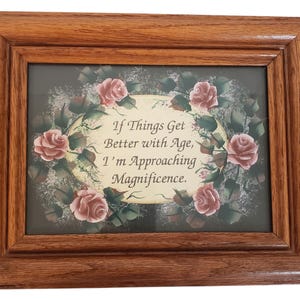 Op de afbeelding: Een rechthoekige houten lijst met een print met een crèmekleurig ovaal midden met de tekst "If Things Get Better with Age, I'm Approaching Magnificence." De tekst is omgeven door een decoratieve rand van roze rozen en groene bladeren.