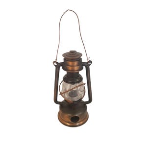 May include: A portable lantern with a metallic bronze finish, featuring a central clear glass globe protected by a wire cage. The lantern includes a top handle for carrying and a circular base with a small opening, showcasing a classic design.