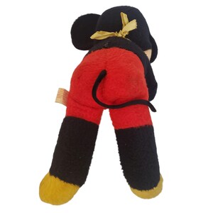 Op de afbeelding: Vintage Mickey Mouse pluche speelgoed, met een zwarte kop en oren, rode broek, zwarte benen en gele voeten. Het pluche heeft een zwarte staart en een gele strik op het hoofd. De achterkant van het speelgoed.