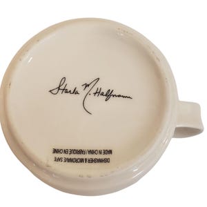 May include: Underside of a white ceramic mug with a handle. The mug features a signature in black ink and the text "DISHWASHER & MICROWAVE SAFE MADE IN CHINA / FABRIQUE EN CHINE".