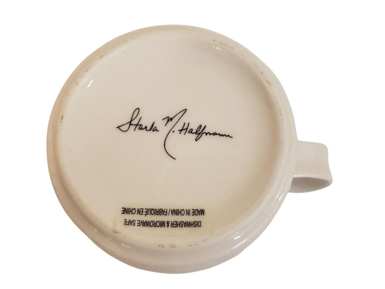 May include: Underside of a white ceramic mug with a handle. The mug features a signature in black ink and the text "DISHWASHER & MICROWAVE SAFE MADE IN CHINA / FABRIQUE EN CHINE".