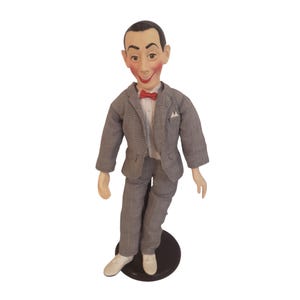 Puede incluir: Muñeco vintage de un hombre con traje gris, camisa blanca y pajarita roja. El muñeco tiene una cara pintada con una amplia sonrisa y está sobre una base negra. El traje tiene un sutil estampado de cuadros.