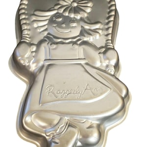 May include: Silver-colored Raggedy Ann cake pan with the words "Happy Birthday" at the top. The pan is shaped like Raggedy Ann, with her signature dress and smiling face. The words "Raggedy Ann" are written on the dress.