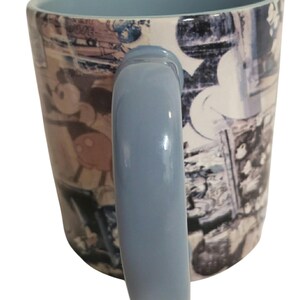 May include: A ceramic mug with a light blue handle and interior. The exterior features a collage of images, including a cartoon character. The mug is white with blue and brown accents.