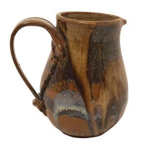 May include: A handmade ceramic pitcher with a curved handle and a spout. The pitcher features a blend of brown, tan, and dark hues, with a glossy finish. The pottery has a unique, organic shape, suitable for serving beverages.
