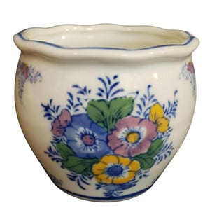 Puede incluir: Una maceta de cerámica blanca con borde festoneado y una línea azul pintada a lo largo del borde. El exterior presenta un motivo floral pintado a mano en tonos azul, amarillo, verde y morado, con delicados detalles de follaje azul.