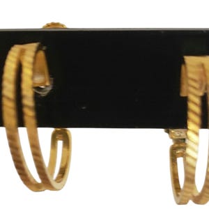 Puede incluir: Un par de pendientes de aro dorados. Cada pendiente presenta un diseño de doble banda con un patrón texturizado y acanalado. Los pendientes se exhiben sobre un fondo negro, mostrando su brillo metálico y sus intrincados detalles.