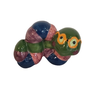 Op de afbeelding: Een keramieken wormfiguur met een glanzende afwerking en gesegmenteerde lichaamsdelen in donkerblauw, gedempt roze en bosgroen. De kop is groen met geschilderde oranje omlijnde brillen en kleine zwarte ogen, wat een kleurrijk beeldje vormt.