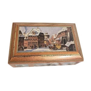 Puede incluir: Una caja rectangular de madera con tapa con bisagras. La tapa presenta una pintura enmarcada de una escena de un pueblo europeo nevado con edificios, un carruaje tirado por caballos y figuras. La caja tiene un borde decorativo.