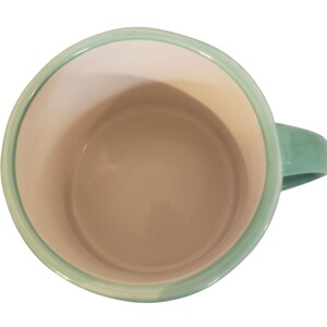 May include: A ceramic mug with a light green handle and rim. The interior is a light beige color. The mug is viewed from above, showcasing its simple, clean design. The mug is suitable for hot or cold beverages.