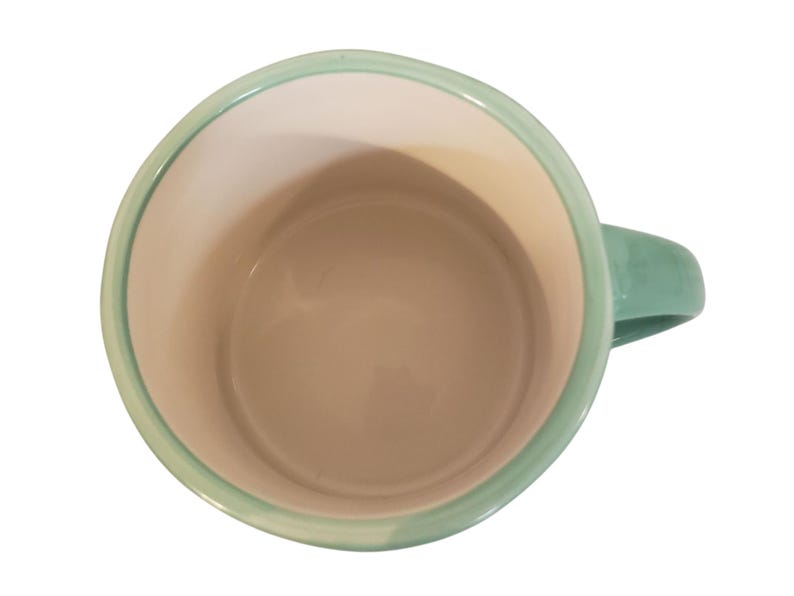 May include: A ceramic mug with a light green handle and rim. The interior is a light beige color. The mug is viewed from above, showcasing its simple, clean design. The mug is suitable for hot or cold beverages.