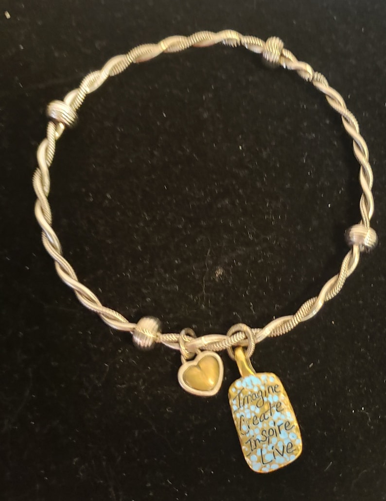 Puede incluir: Pulsera de brazalete plateada con un dise&ntilde;o de cuerda trenzada. La pulsera presenta un dije de coraz&oacute;n dorado y un dije rectangular con las palabras "Imagine Create Inspire Live" en negro y azul.