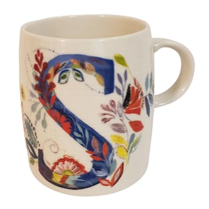 May include: Cream-colored ceramic mug featuring a large, floral-decorated letter "S" in blue. The design includes red, yellow, and green floral accents. The mug has a curved handle and a smooth, glossy finish, perfect for coffee or tea.