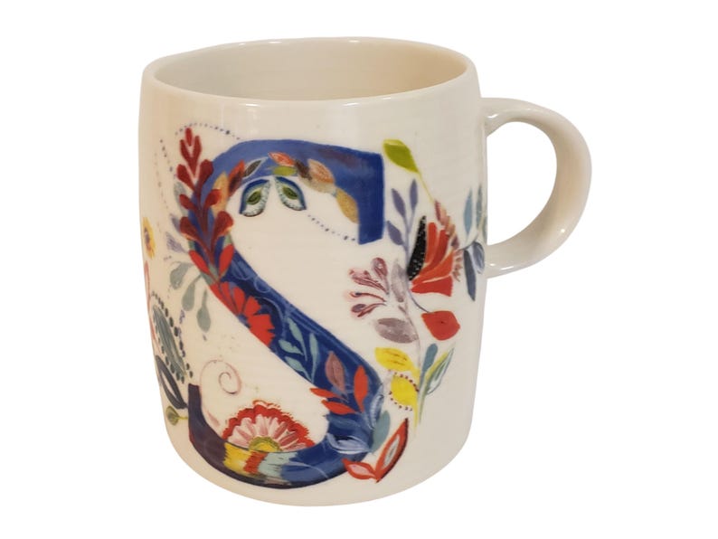 May include: Cream-colored ceramic mug featuring a large, floral-decorated letter "S" in blue. The design includes red, yellow, and green floral accents. The mug has a curved handle and a smooth, glossy finish, perfect for coffee or tea.