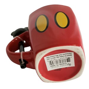 May include: A red ceramic mug featuring two yellow oval shapes with black outlines on the front. The mug has a black handle and a white base with a product label containing a barcode and text reading Jerry Leigh Orlando, FL 32837, Made in China.