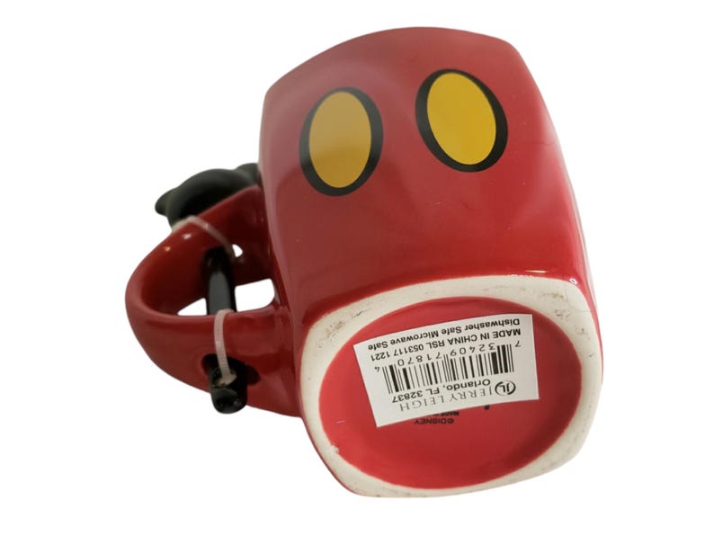 May include: A red ceramic mug featuring two yellow oval shapes with black outlines on the front. The mug has a black handle and a white base with a product label containing a barcode and text reading Jerry Leigh Orlando, FL 32837, Made in China.