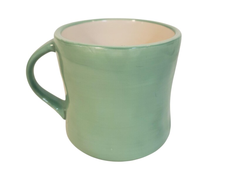 May include: A light green ceramic mug with a curved body and a handle. The interior of the mug is white. The mug is designed with a smooth, glossy finish. This mug is suitable for hot or cold beverages.